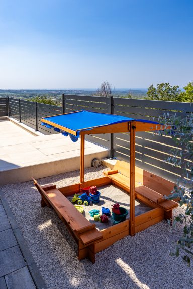 Sandkasten aus Holz mit blauem Sonnendach und Spielzeug, angebunden an die Terrasse, Aussicht im Hintergrund.